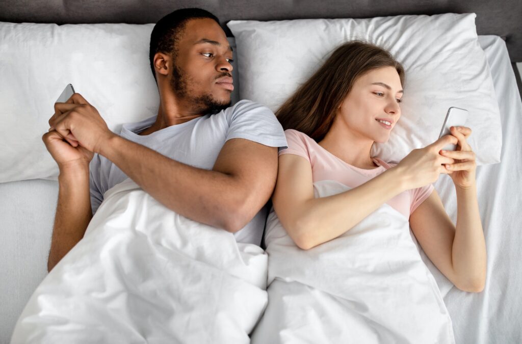 Young interracial couple lying in bed stuck in smartphones, playing online games, browsing social